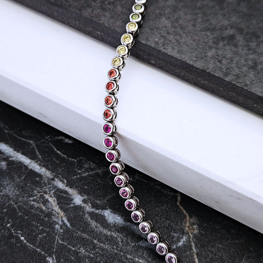 A silver-toned chakra bracelet with multiple colorful gemstones, draped diagonally across a white book on a dark marble surface.