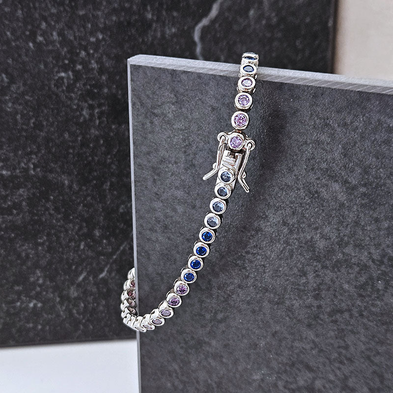 A silver-toned tennis bracelet with alternating round blue and purple gemstones, draped over a dark gray stone display block.
