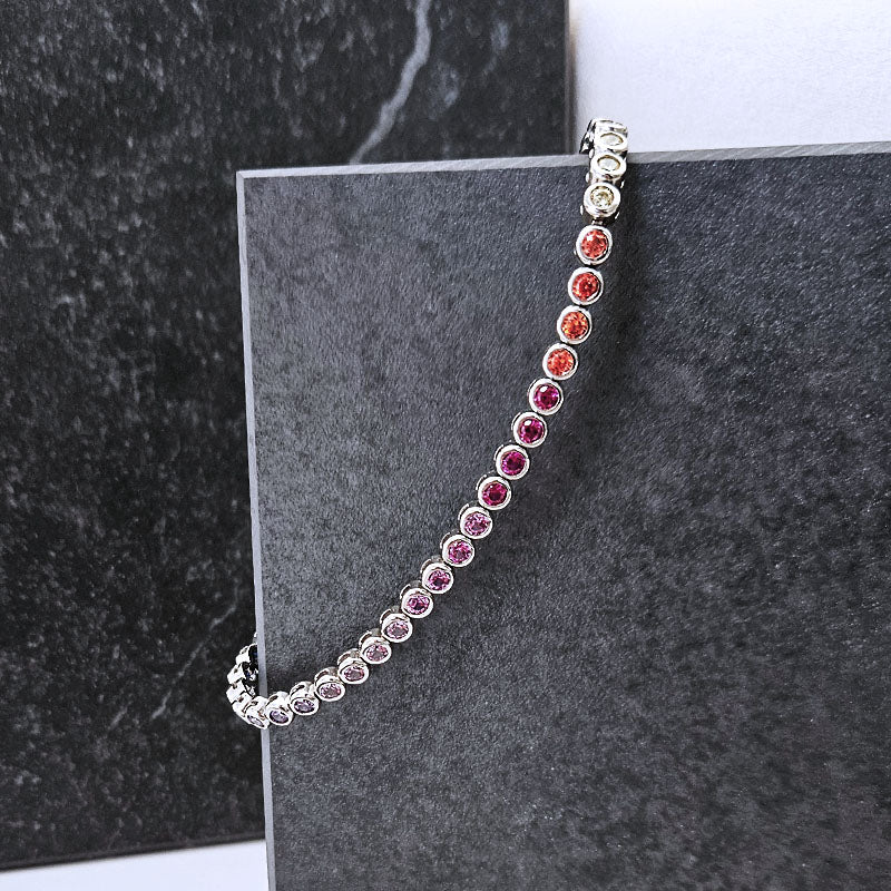 A silver tennis bracelet with round gemstones in a gradient of colors from clear to orange, pink, and purple, displayed on a dark gray textured block.