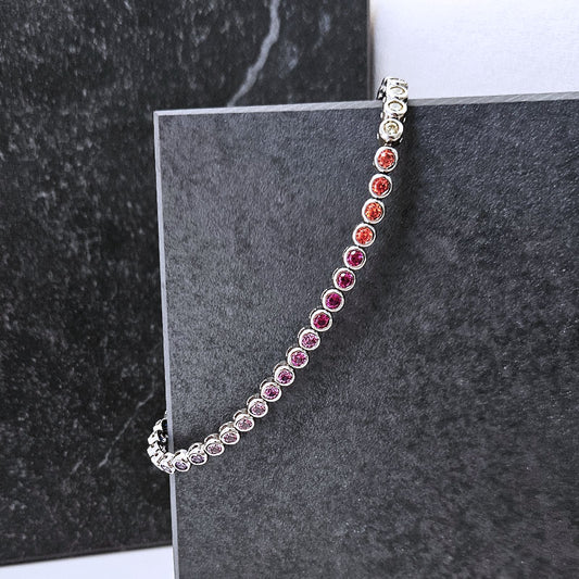 A silver tennis bracelet with round gemstones in a gradient of colors from clear to orange, pink, and purple, displayed on a dark gray textured block.