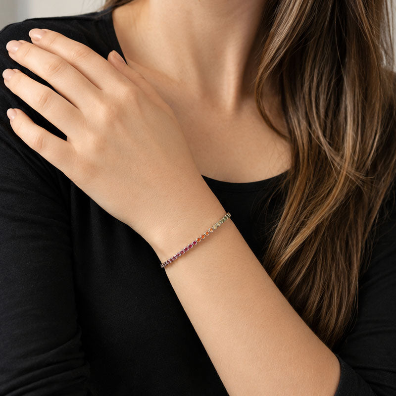 A close-up of a woman wearing a delicate rainbow-colored chain bracelet on her wrist, paired with a black long-sleeve top.