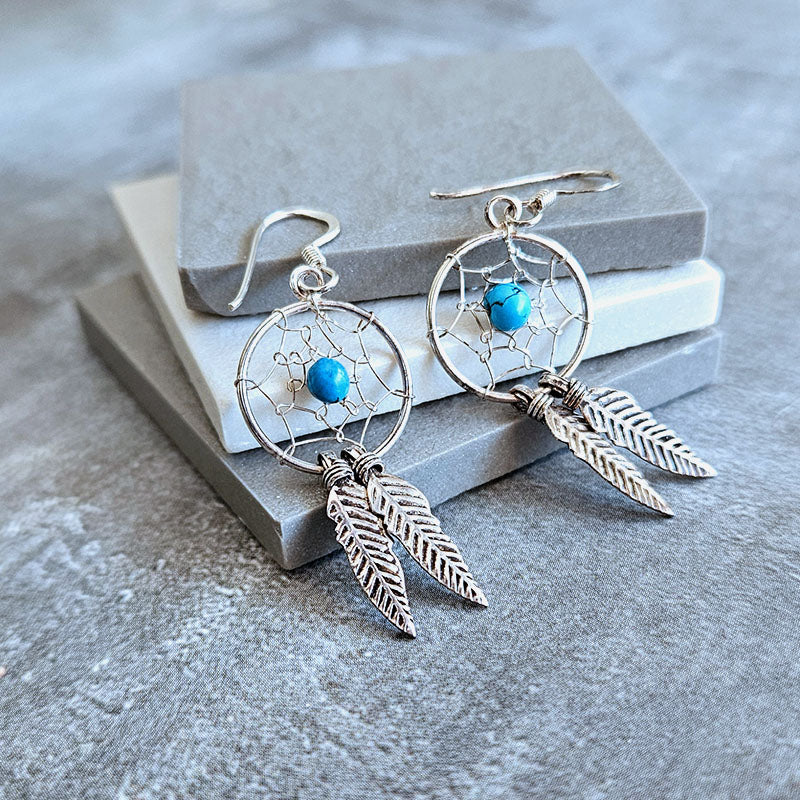 Silver dreamcatcher earrings with blue stones and feathers on a gray surface.
