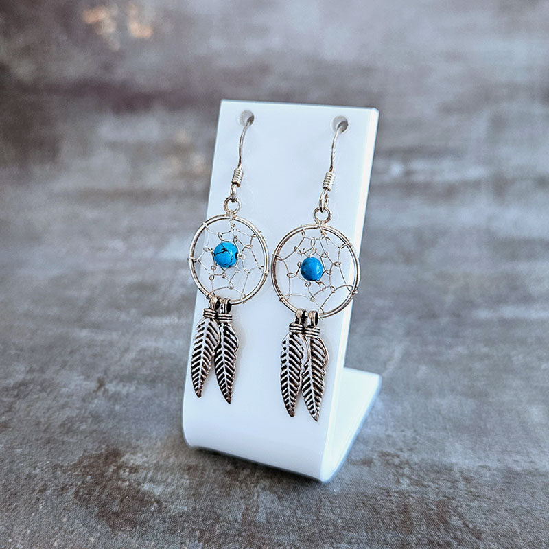 Dreamcatcher earrings with blue stones and feather designs on a white stand against a gray background.