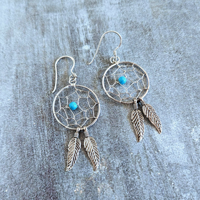 Silver dreamcatcher earrings with turquoise stones on a textured gray surface