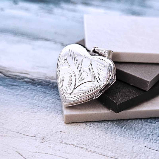 Heart-shaped silver locket with engraved design on a textured surface
