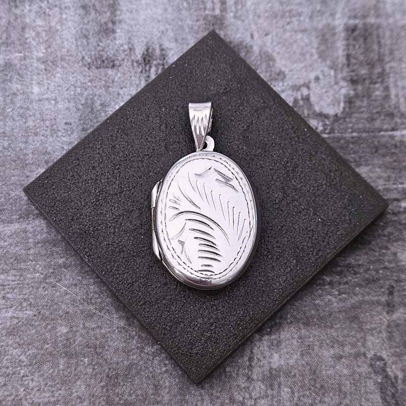 Silver locket with engraved design on a black square pad against a textured gray background