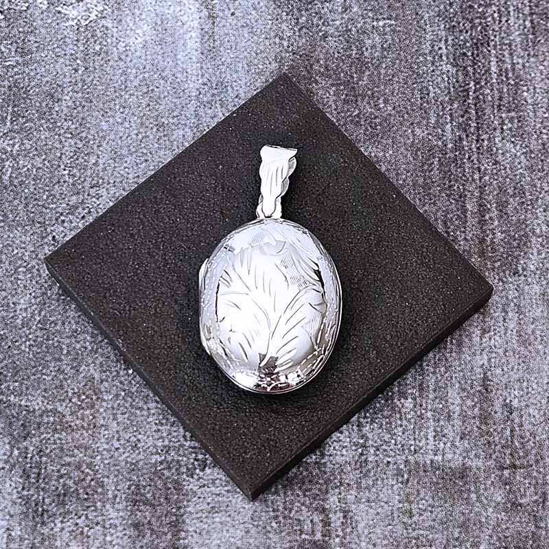 Silver locket with engraved design on a textured black stand against a gray background