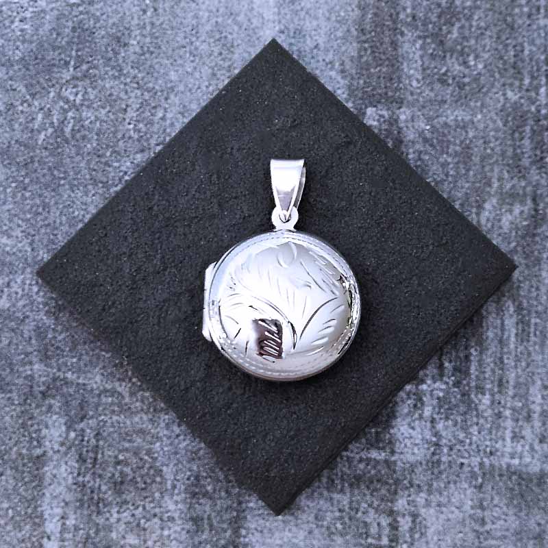 Silver locket with engraved design on a black square cushion against a textured gray background