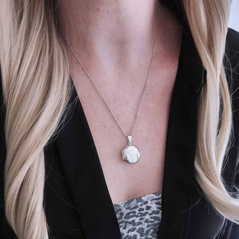 Person wearing a silver necklace with a round pendant, black blazer, and patterned top.