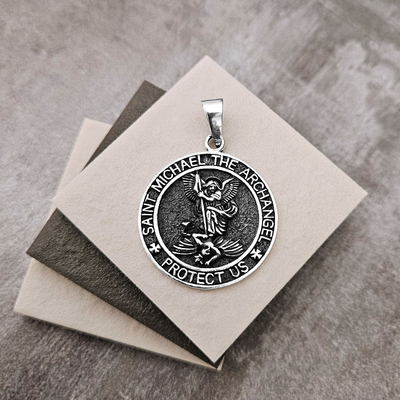 Silver pendant with 'Saint Michael the Archangel Protect Us' text on a gray surface