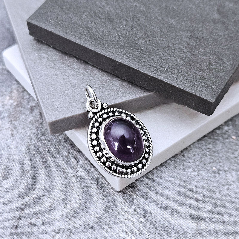 An oval pendant featuring a deep purple cabochon gemstone surrounded by a silver-tone beaded border, displayed on stacked gray stone blocks.
