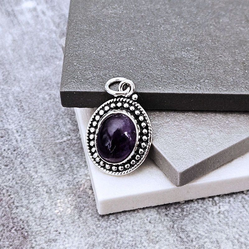 An oval pendant featuring a deep purple amethyst gemstone in a silver-toned bezel setting with a beaded border, displayed on stacked gray stone blocks.