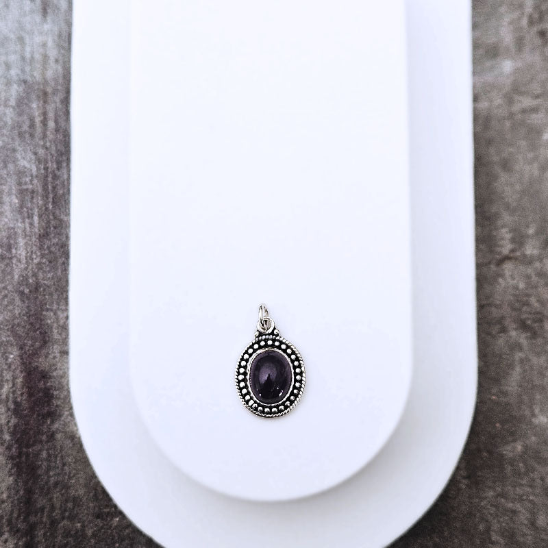 A silver-toned oval pendant with a dark purple amethyst center stone, displayed on a white jewelry stand against a textured gray background.