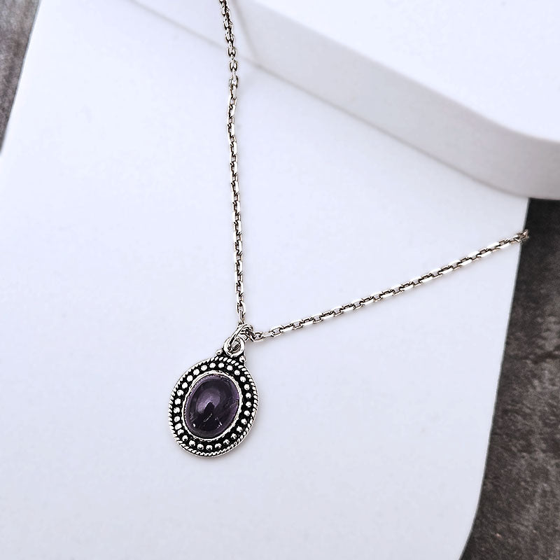 A silver-toned chain necklace with an oval pendant featuring a dark purple cabochon stone surrounded by a beaded silver border, displayed on a white surface.