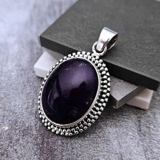 An oval pendant featuring a deep purple cabochon gemstone in a silver bezel setting with a beaded border, displayed on a textured gray surface with dark gray and white blocks.