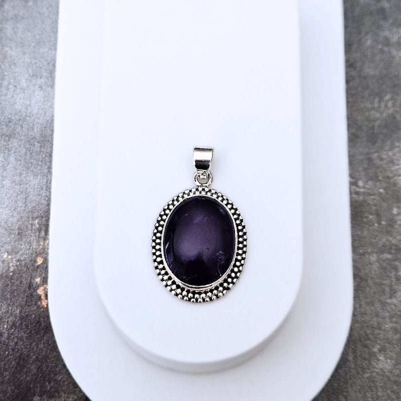An oval pendant with a dark purple cabochon stone and a silver beaded border, displayed on a white jewelry stand.