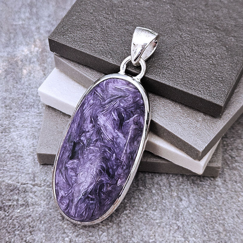An oval silver pendant featuring a marbled purple stone, displayed on a stack of gray and white blocks against a textured gray background.