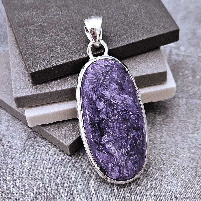 An oval pendant featuring a marbled purple stone in a silver bezel setting, displayed on stacked gray and white blocks.
