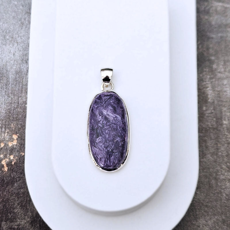 An oval pendant featuring a textured purple stone with silver bezel and bail, displayed on a white stand against a gray textured background.