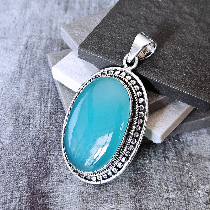 An oval chalcedony pendant with a silver-toned filigree border, displayed on stacked gray stone blocks.