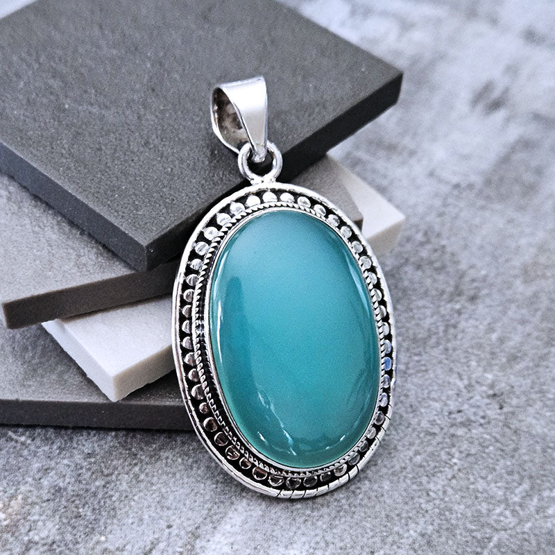 An oval pendant featuring a smooth, glossy chalcedony stone in a detailed silver-toned bezel setting, displayed on stacked dark gray and white stone slabs against a textured gray background.