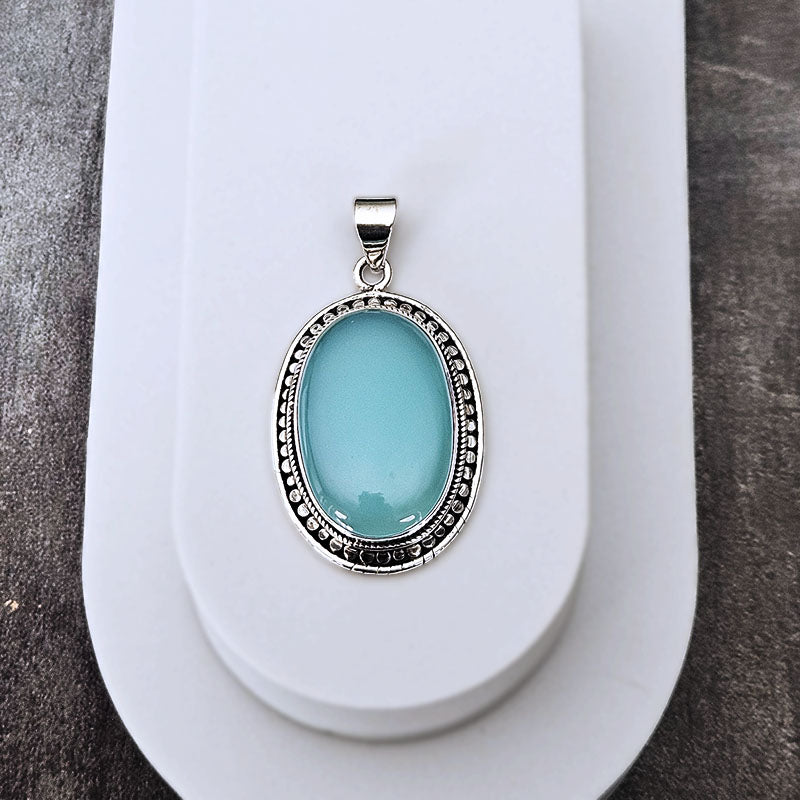 An oval pendant featuring a smooth light blue-green stone in a silver-toned bezel setting, displayed on a white stand against a dark textured background.