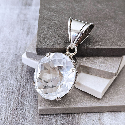 A silver pendant with a large, faceted clear oval gemstone, displayed on stacked gray and white boxes.