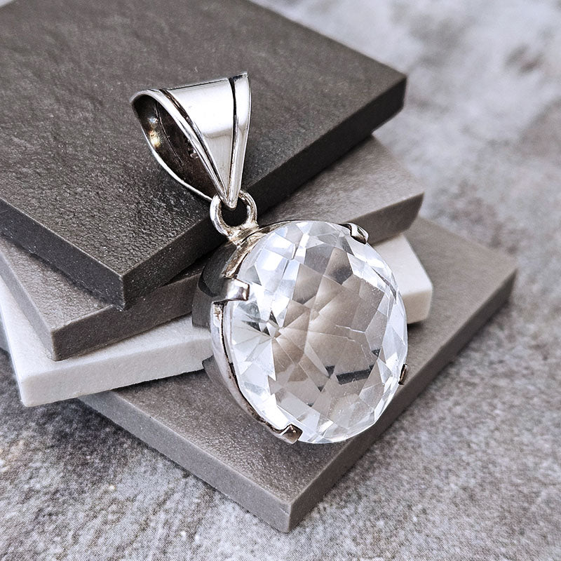 A silver pendant with a large, round, faceted clear gemstone, displayed on stacked gray and white stone tiles.