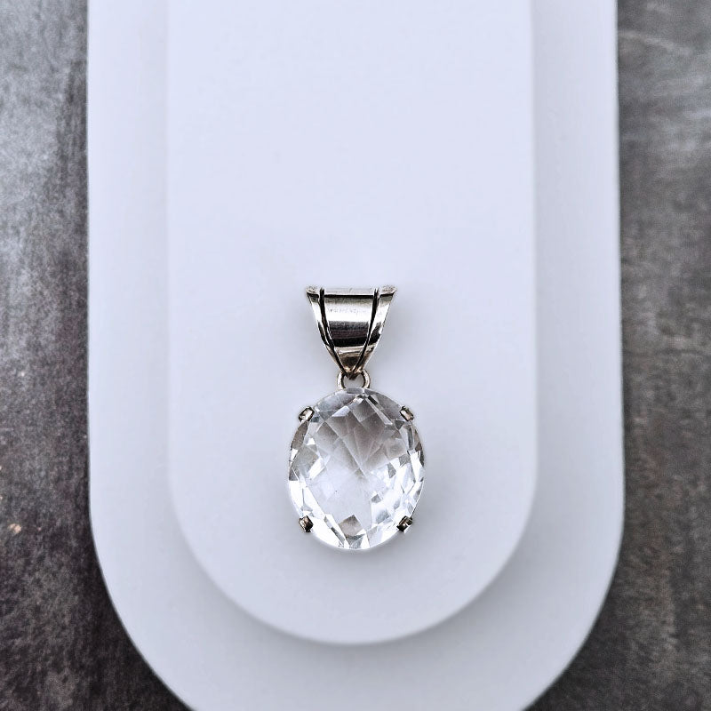 A faceted clear oval gemstone pendant in a silver-toned setting, displayed on a white curved stand against a dark gray textured background.