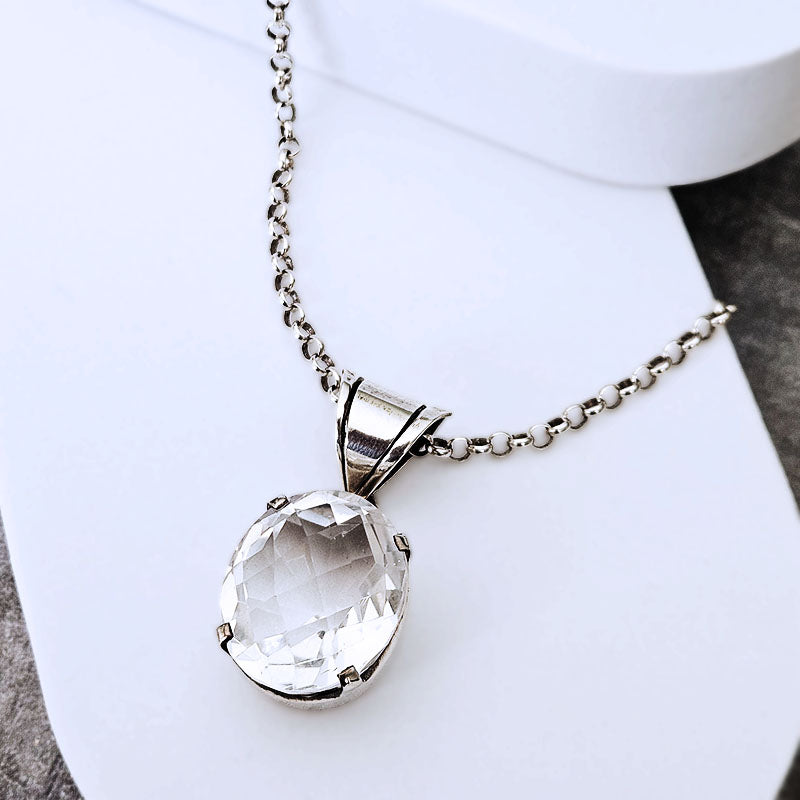 A silver-toned chain necklace with a faceted clear oval pendant, displayed on a white surface.