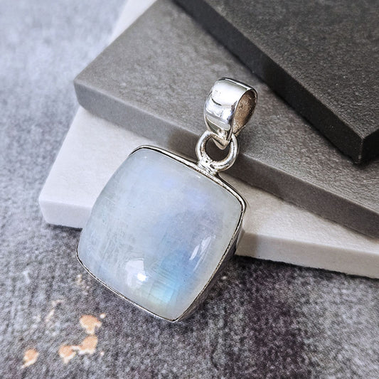 A square pendant featuring a milky white iridescent stone with soft blue-green flashes, set in a polished silver bezel with a matching silver bail, displayed on stacked gray and white stone blocks.