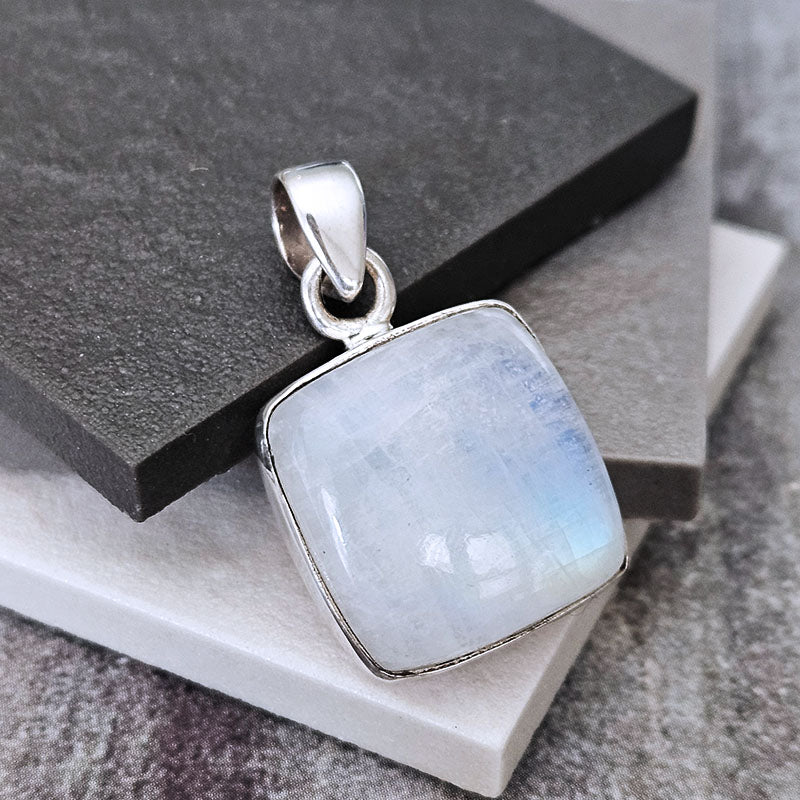 A square silver pendant featuring a milky white iridescent gemstone with subtle blue and green flashes, set in a polished silver bezel and bail, resting on stacked dark gray and white stone blocks.