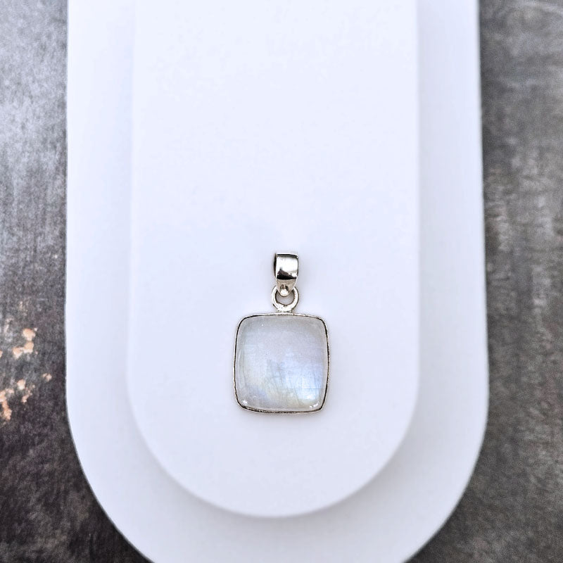 A square pendant with a milky white iridescent stone in a silver bezel setting, displayed on a white stand against a dark textured background.