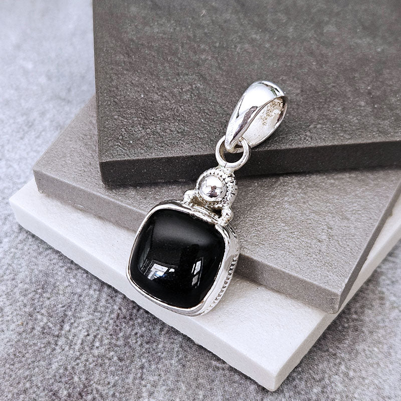 A silver pendant with a square black onyx stone, displayed on stacked gray and white stone blocks.