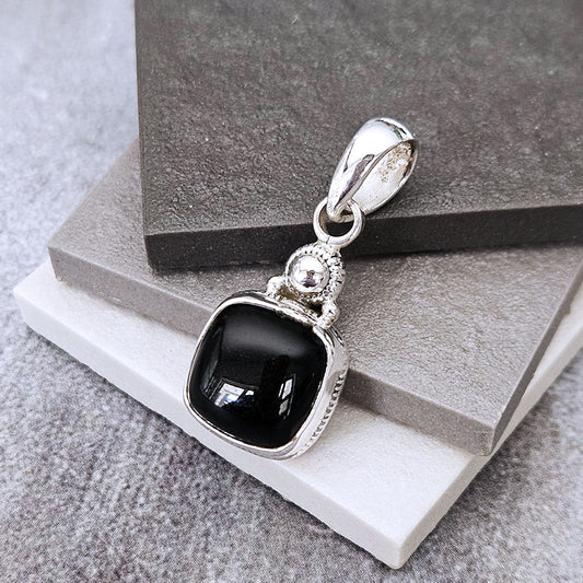 A silver pendant with a square black onyx stone, displayed on stacked gray and white stone blocks.