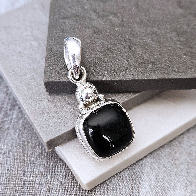 A silver pendant with a square black onyx stone, resting on stacked gray and white textured stone tiles.