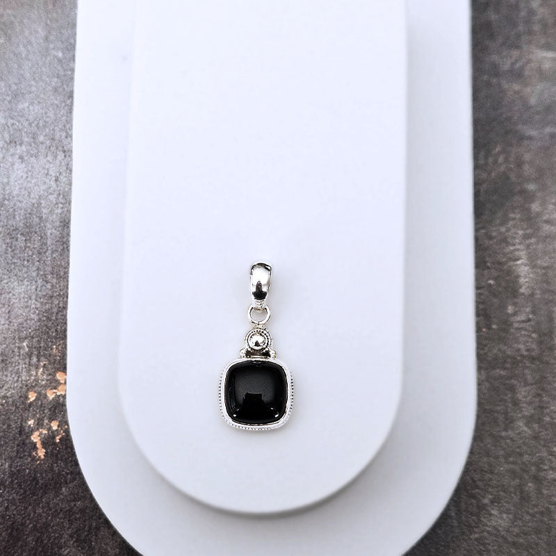 A silver-toned pendant with a square black gemstone and a small clear accent stone, displayed on a white jewelry stand against a dark textured background.
