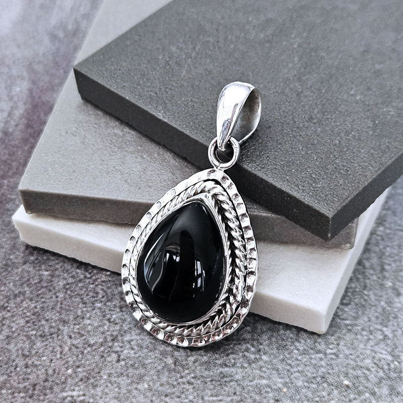 A silver teardrop pendant with a polished black onyx center, resting on stacked gray and white display blocks.