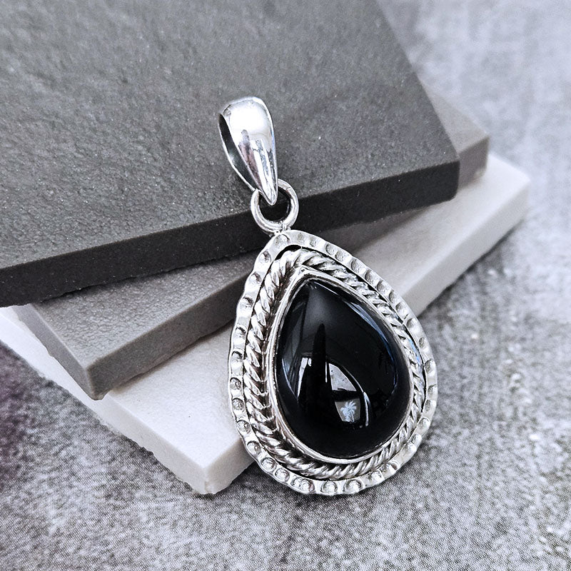 A silver teardrop pendant with a polished black onyx stone, resting on stacked gray and white stone slabs.