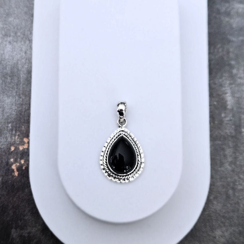 A silver teardrop pendant with a textured silver border and a glossy black teardrop gemstone, displayed on a white jewelry stand against a dark gray textured background.