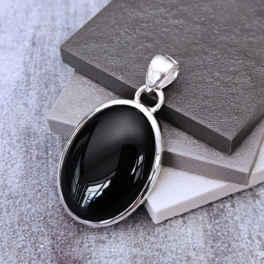 A silver oval pendant with a glossy black cabochon gemstone, displayed on stacked gray and white blocks against a textured gray background.