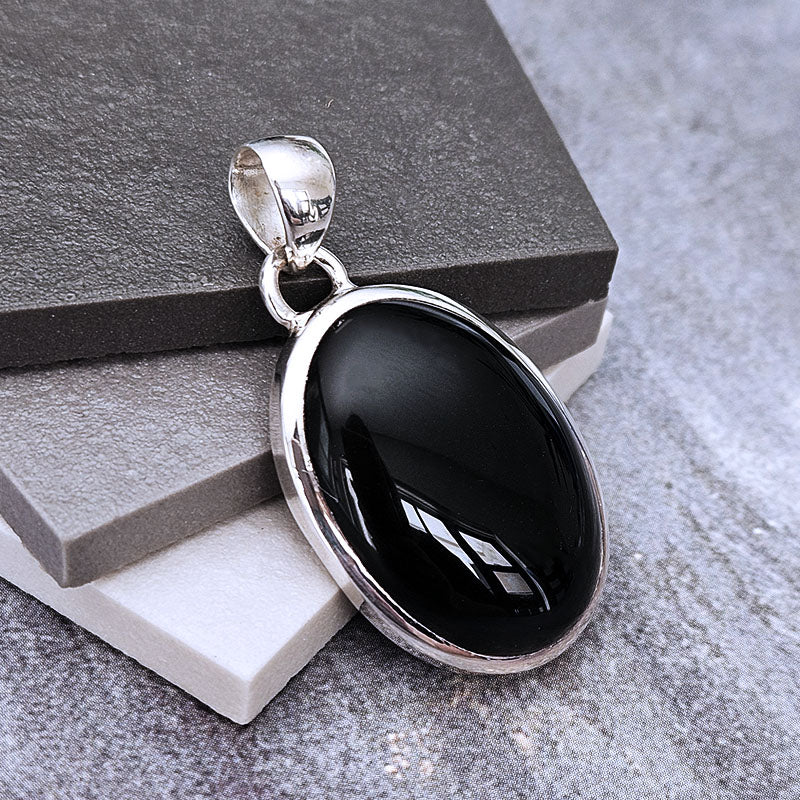 A silver oval pendant with a glossy black cabochon stone, displayed on stacked gray and white stone blocks.