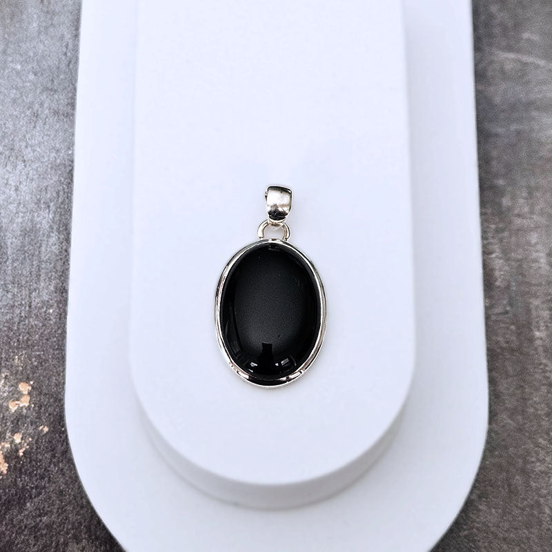 A silver oval pendant with a smooth, polished black stone, displayed on a white jewelry stand against a textured gray background.