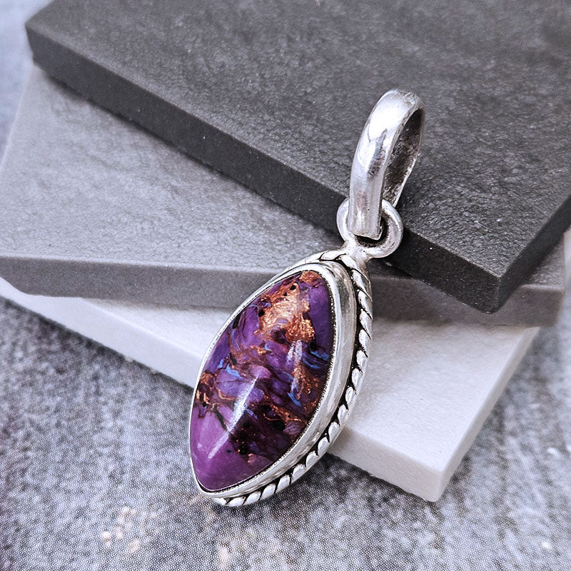 A silver pendant featuring a teardrop-shaped purple stone with coppery metallic inclusions, displayed on a gray and white stone block background.
