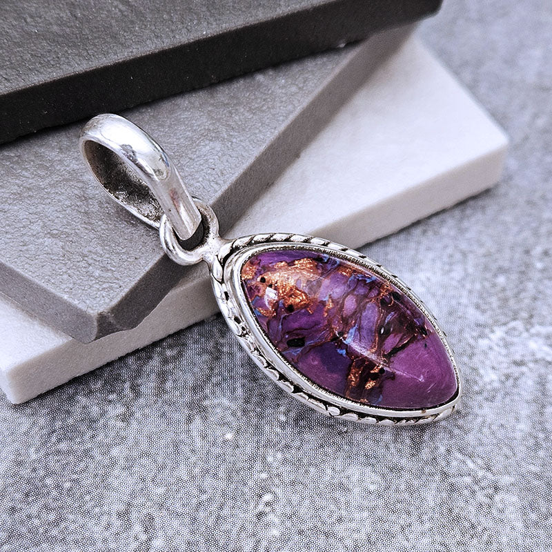 A silver pendant featuring an oval purple stone with brown inclusions, displayed on a gray textured surface with a white and gray geometric block.