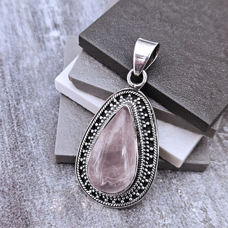 A teardrop-shaped pendant featuring a faceted light pink gemstone surrounded by an ornate silver filigree border, displayed on stacked dark gray and white stone slabs against a textured gray background.