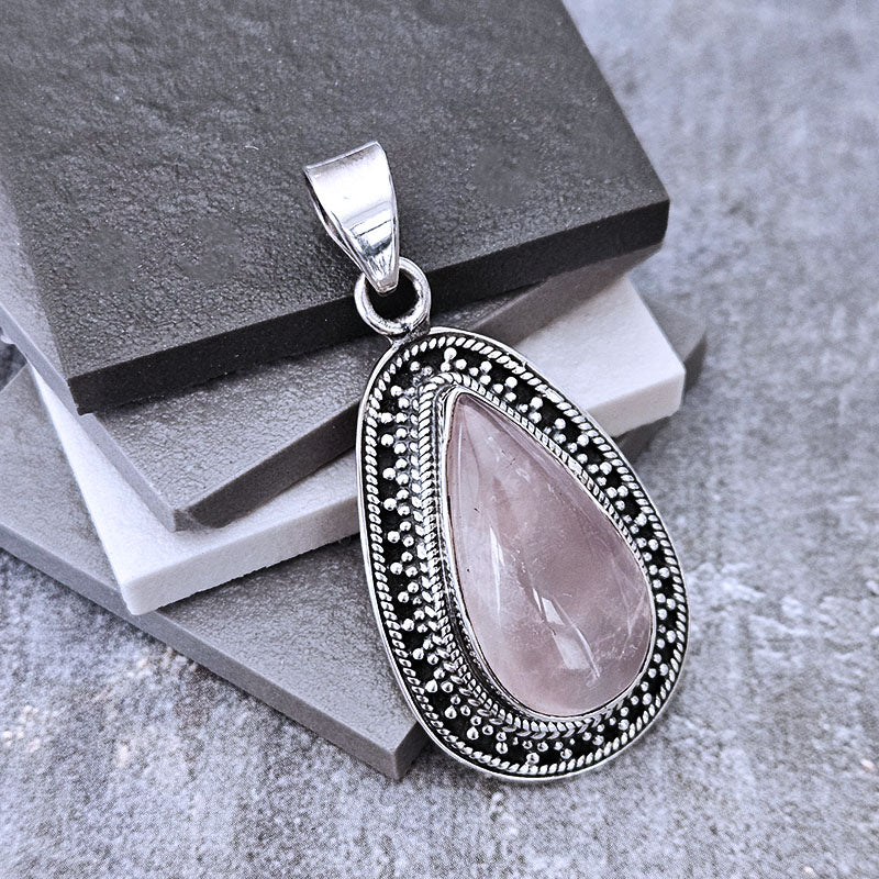 A silver teardrop pendant featuring a light pink rose quartz stone, set in an ornate silver bezel with black oxidized details, displayed on stacked dark gray stone blocks.