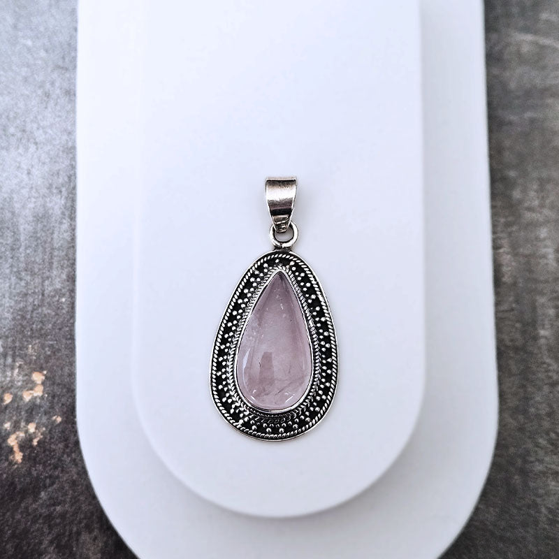 A teardrop-shaped pendant featuring a light pink rose quartz center stone surrounded by a silver-tone border with black oxidized details, displayed on a white stand.