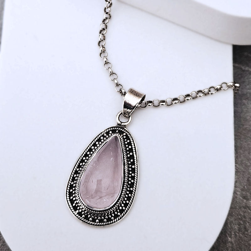 A silver-toned teardrop pendant necklace with a faceted light pink gemstone, set in an ornate silver bezel with a delicate chain, displayed on a white curved stand.