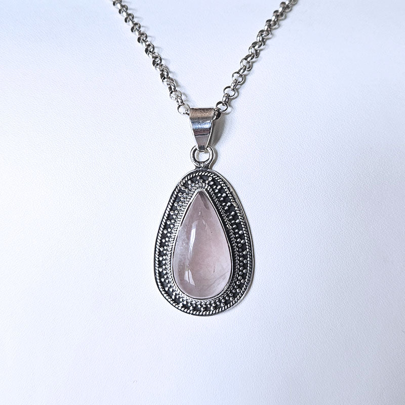 A silver teardrop pendant necklace featuring a pale pink rose quartz center stone surrounded by a silver filigree border, suspended from a silver chain against a plain white background.